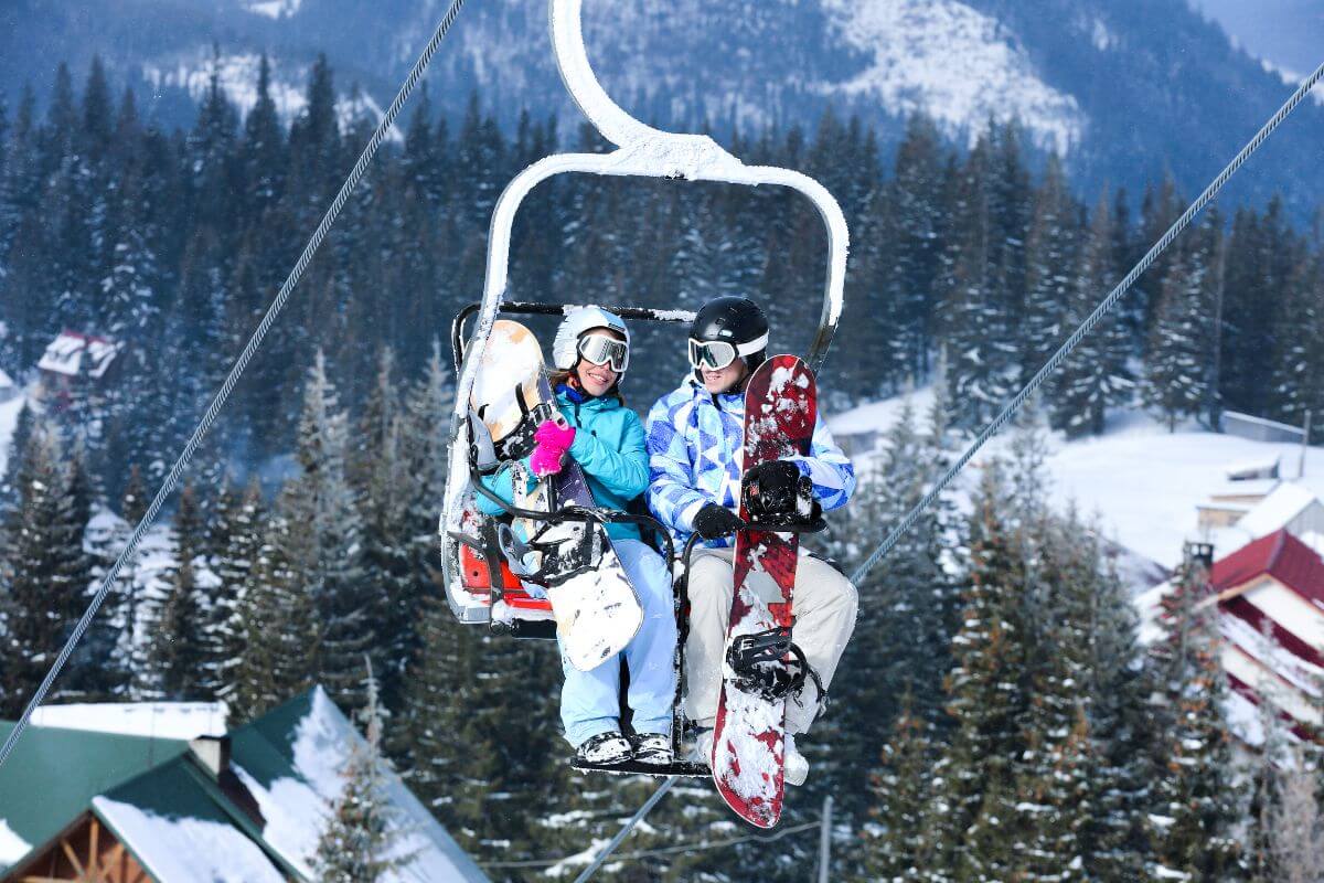 Two people sitting on a ski lift with their snowboards.