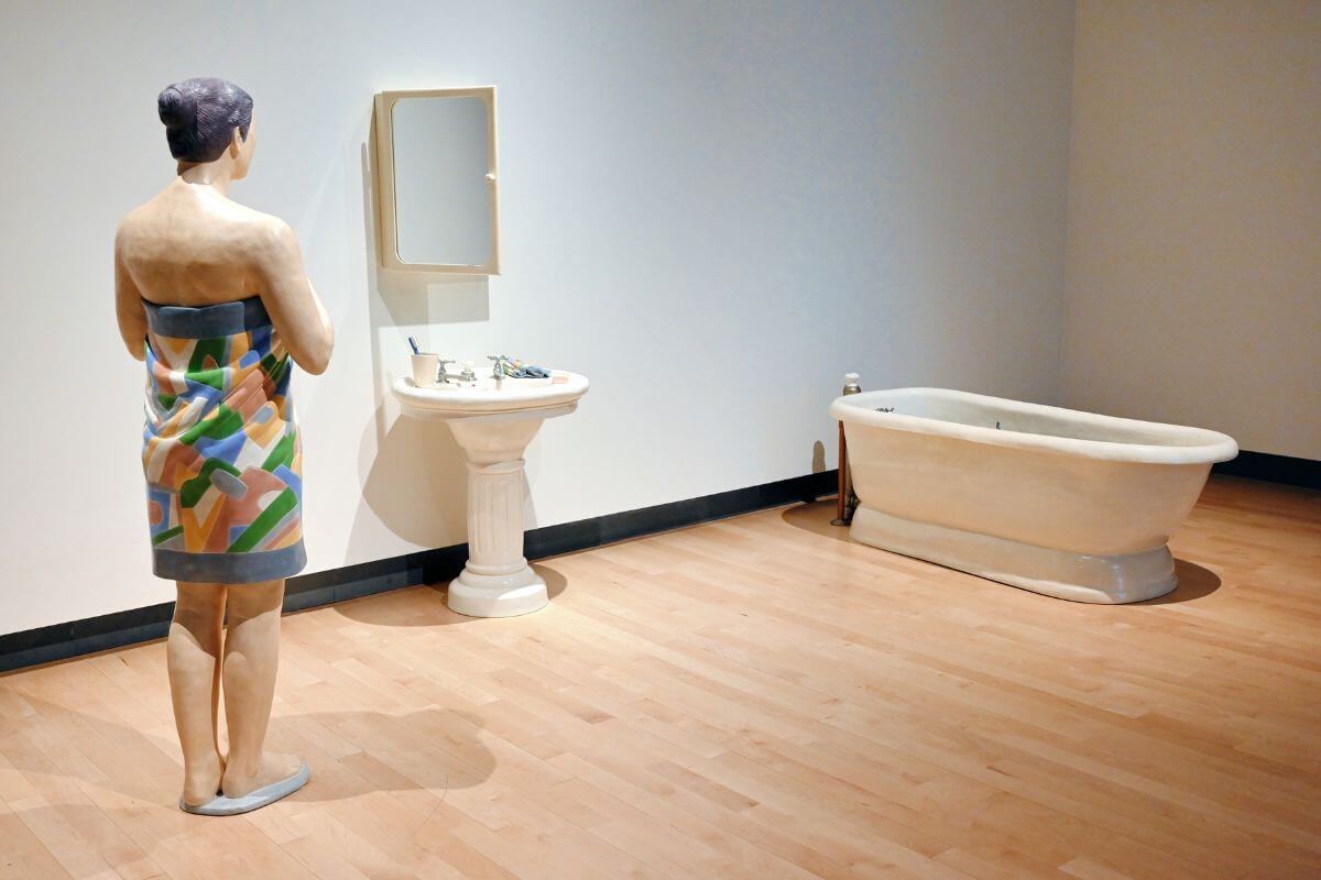In a Montana gallery, an art exhibit highlights a sculpture depicting a person in a towel within a bathroom setting, complete with sculpted sink, mirror, and bathtub.