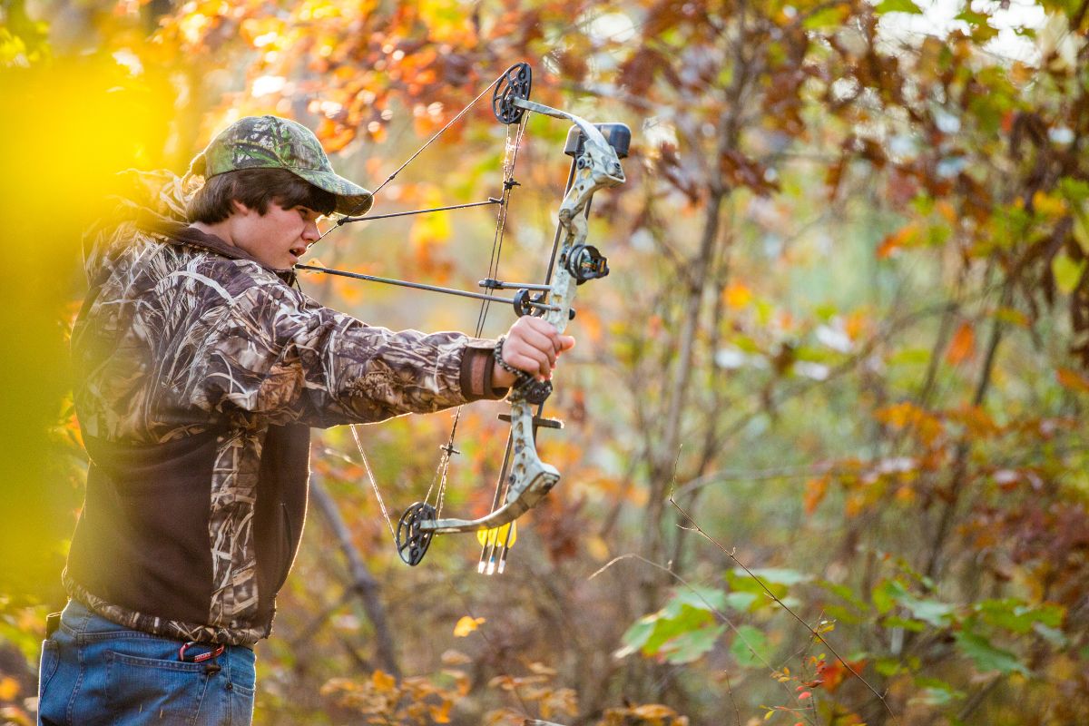 A young man in camouflage aims a compound bow in an autumn forest near Pintler Waterfall, Montana, with golden leaves in the background.