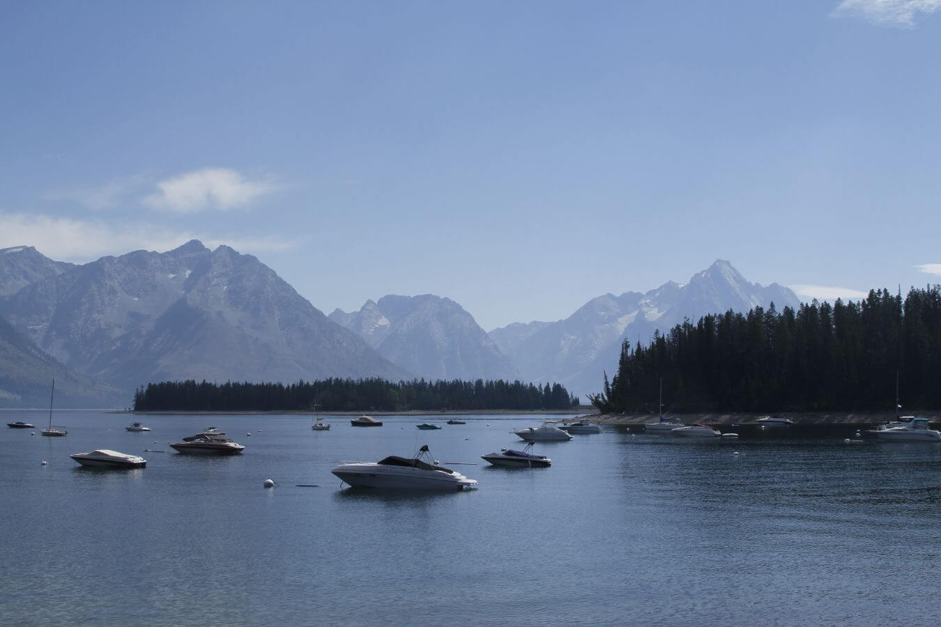 A peaceful lake in Montana with multiple boats floating on its surface, surrounded by majestic, forest-covered mountains under a clear blue sky.