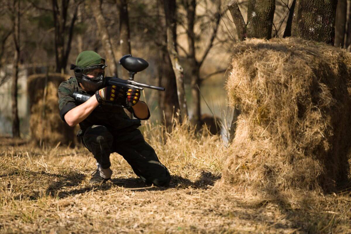 A man with a paintball gun aims at a target from behind a bale of hay in a Montana paintball field.